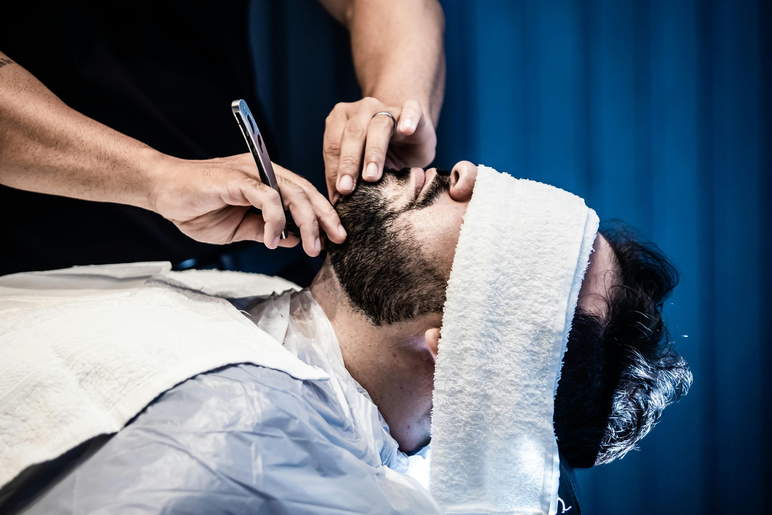 An adult man receiving a precise beard trim in a barbershop with a warm towel treatment.